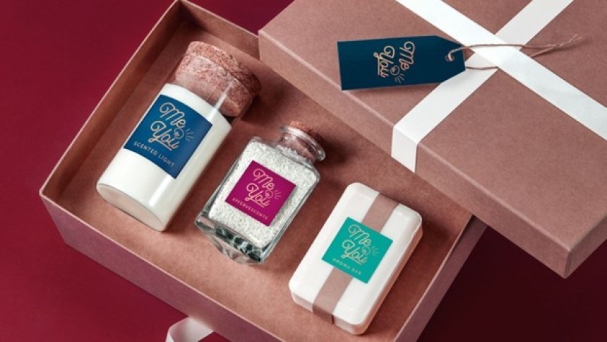An open gift set in a brown box containing three spa products: a scented candle, bath salts, and a bar of soap. Each product has a label featuring the “Me & You” logo.