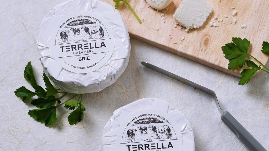 Two round wheels of brie cheese are displayed with white paper labels for "Terella Creamery." In the background, a wooden board holds cut pieces of brie next to a cheese knife and fresh parsley.
