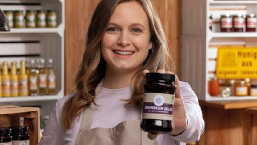 A friendly young woman with light brown hair, wearing a beige apron, smiles warmly as she presents a jar of "Brombeer Gelee" (blackberry jelly) to the camera. She is in a rustic shop with wooden shelves stocked with other jars of preserves.