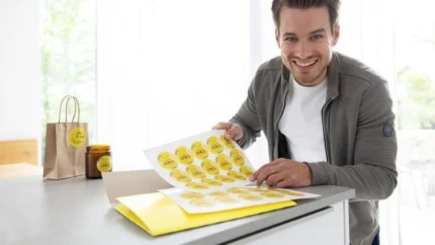 A smiling man holds a sheet of freshly printed round, yellow labels, with a branded paper bag and jar featuring the same label next to him.