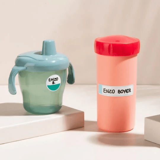 Green transparent plastic cup with spout and handle, and salmon-colored elongated plastic bottle with red cap, on which handwritten stickers are placed.