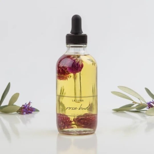 A clear glass dropper bottle of La Luna Rose Bud body oil, filled with a golden oil and whole, dried magenta rosebuds. The bottle is flanked by sprigs of lavender and olive leaves.