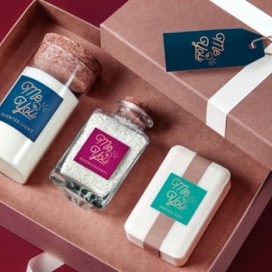 An open gift set in a brown box containing three spa products: a scented candle, bath salts, and a bar of soap. Each product has a label featuring the “Me & You” logo.