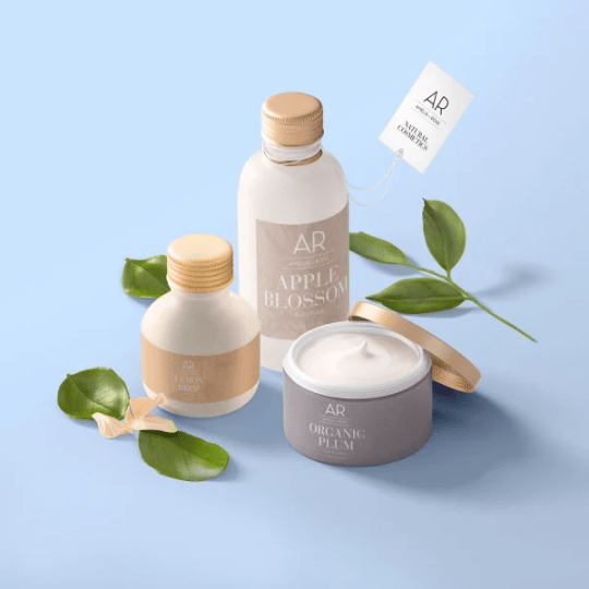 A collection of AR Amelia Rose natural cosmetics on a light blue background, featuring Organic Plum cream in an open grey jar, Lemon Drop lotion, and Apple Blossom Body Polish. The products are styled with fresh green leaves.