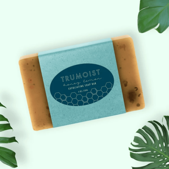 A top-down view of a Trumoist Honey Lemon Exfoliating Soap Bar. The translucent, honey-colored soap bar is wrapped in a blue paper sleeve with a honeycomb design, displayed on a mint green background with tropical leaves in the corners.
