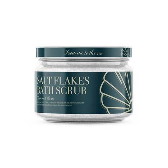 A clear glass jar of "From me to the sea" Salt Flakes Bath Scrub. The jar has a dark teal label and lid, featuring a stylized art deco seashell design.
