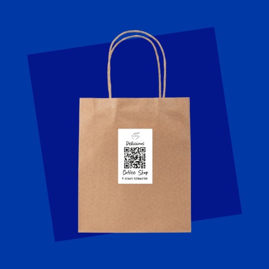 A brown paper bag against a blue background, with a white label applied to its center. The label features the word 'Delicious' next to a coffee cup icon, a QR code, and the text 'Coffee Shop' with a telephone number below.