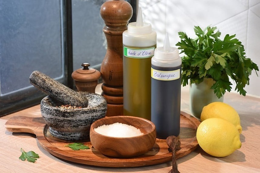 A wooden tray with a stone mortar, a wooden salt shaker, a wooden pepper shaker, two plastic oil bottles, two lemons, and herbs in a bowl.