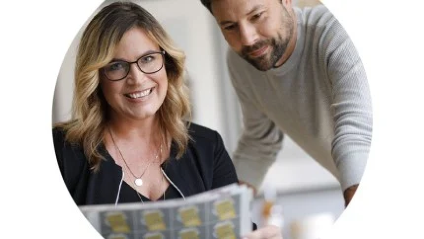 A smiling woman wearing glasses is sitting at a desk, showing a sheet of designs. Behind her stands a man who is watching her work with interest.