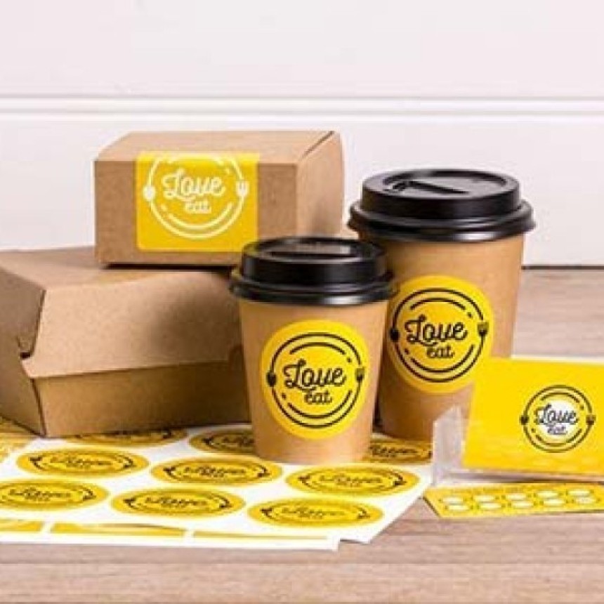 A collection of takeaway packaging, including two coffee cups and a cardboard box, is branded with bright yellow circular "Love eat" logo stickers. Sheets of the unused stickers are displayed in the foreground.