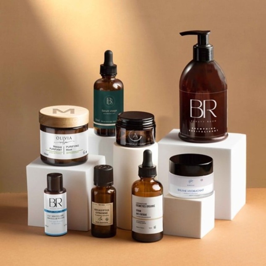 A collection of various skincare products in elegant, mostly amber-colored packaging. The collection includes bottles with pumps and droppers, as well as jars. The products are arranged on white rectangular blocks against a neutral background.