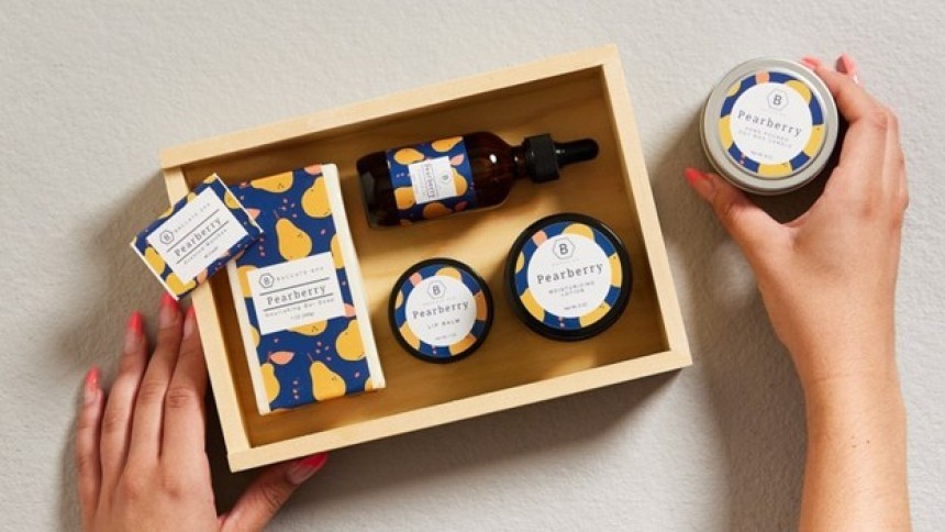 A set of Pearberry cosmetics on a light-colored wooden tray. The packaging features a cohesive, colorful design with a pear motif. A woman’s hands are holding a round metal box containing lotion.
