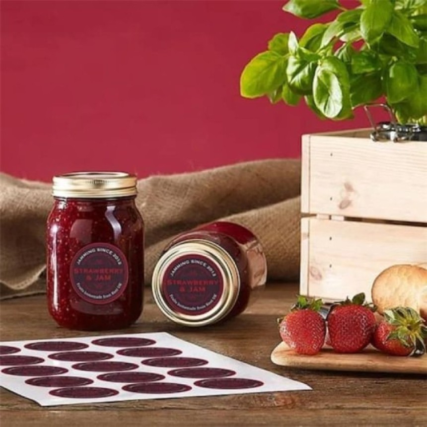 A rustic still life featuring two jars of strawberry jam with gold lids and custom "Strawberry & Jam" labels. One jar stands upright while the other is on its side. The scene is set on a wooden table with burlap, a sheet of the matching labels, fresh strawberries, and a basil plant in a wooden crate.