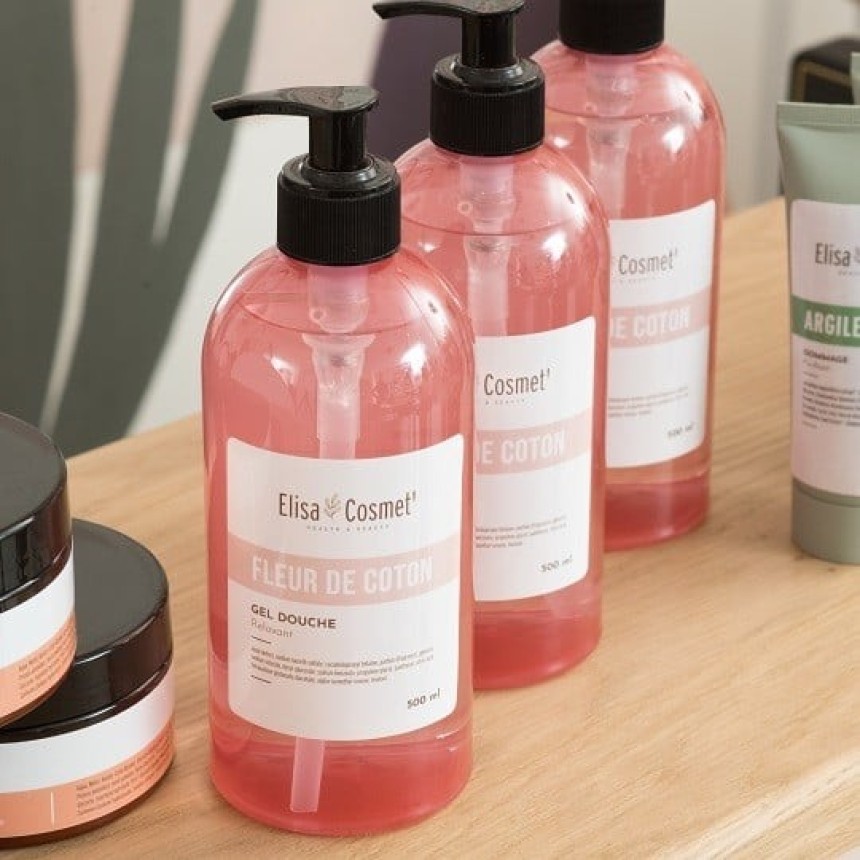 Three clear pump bottles of Elisa Cosmet's "Fleur de Coton" pink shower gel arranged on a light wooden surface, with other skincare products partially visible.