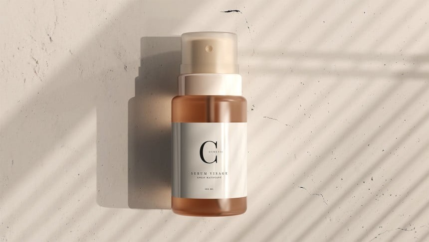A small, amber-colored spray bottle of Genetic Sérum Visage. The bottle is lying on a textured, light-colored surface, with a dramatic shadow of window blinds falling across it.