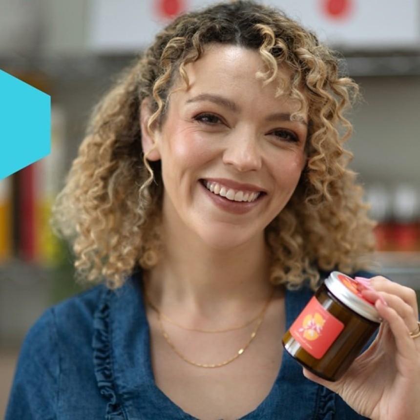 A smiling woman with curly hair presents a small product jar with a colorful label.