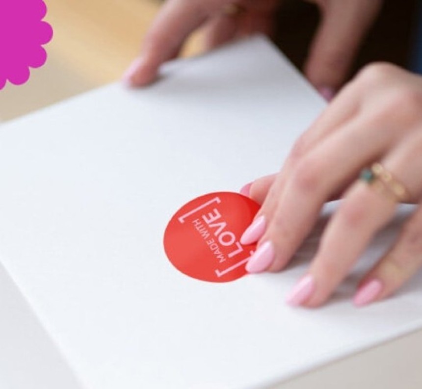 A woman's hands are applying a round, red sticker that says "Made with love" to a white box.